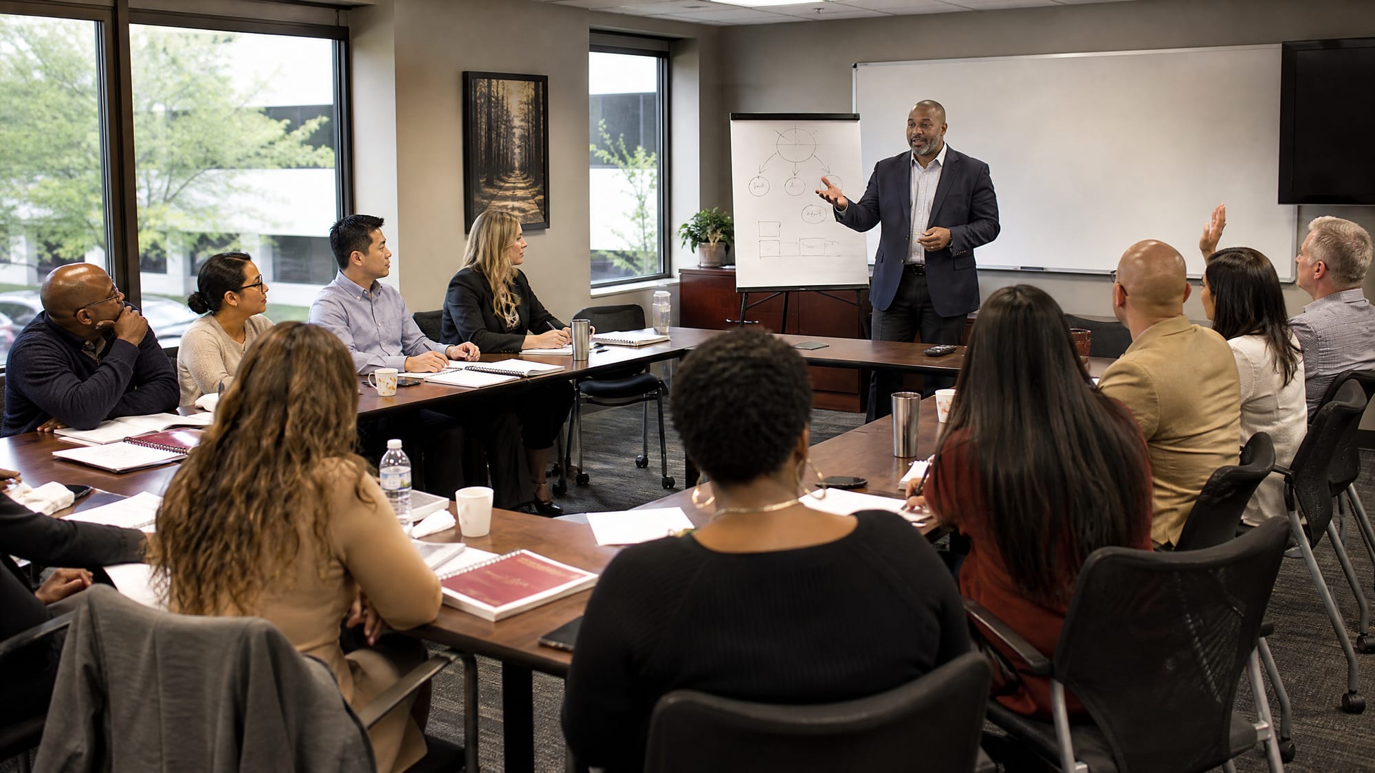 Instructor leading a live management training class at a whiteboard, a dozen participants around U-shaped tables